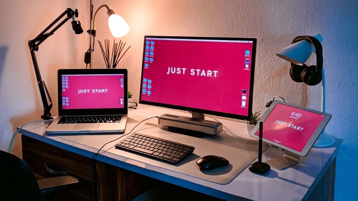 Espacio de trabajo con una MacBook Pro, un monitor y un iPad. Todos muestran en pantalla la frase "Just Start"