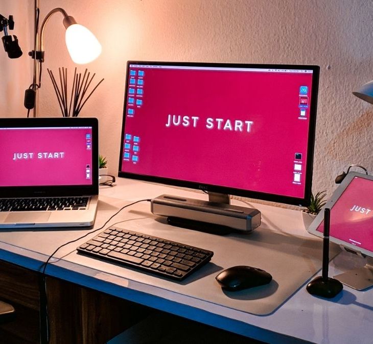 Espacio de trabajo con una MacBook Pro, un monitor y un iPad. Todos muestran en pantalla la frase "Just Start"